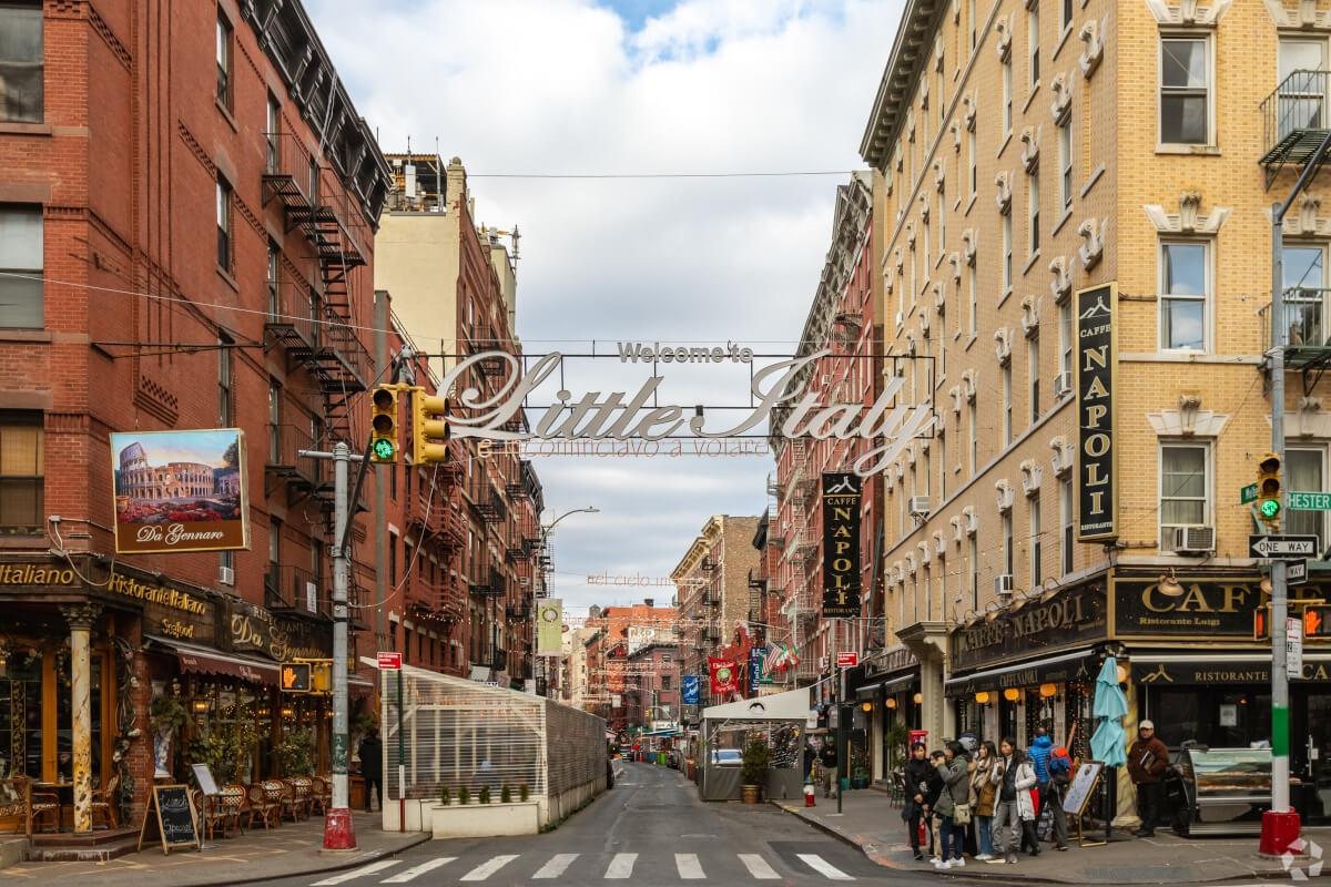A sign reading “Welcome to Little Italy” hangs over Mulberry Street between Italian-style cafés.