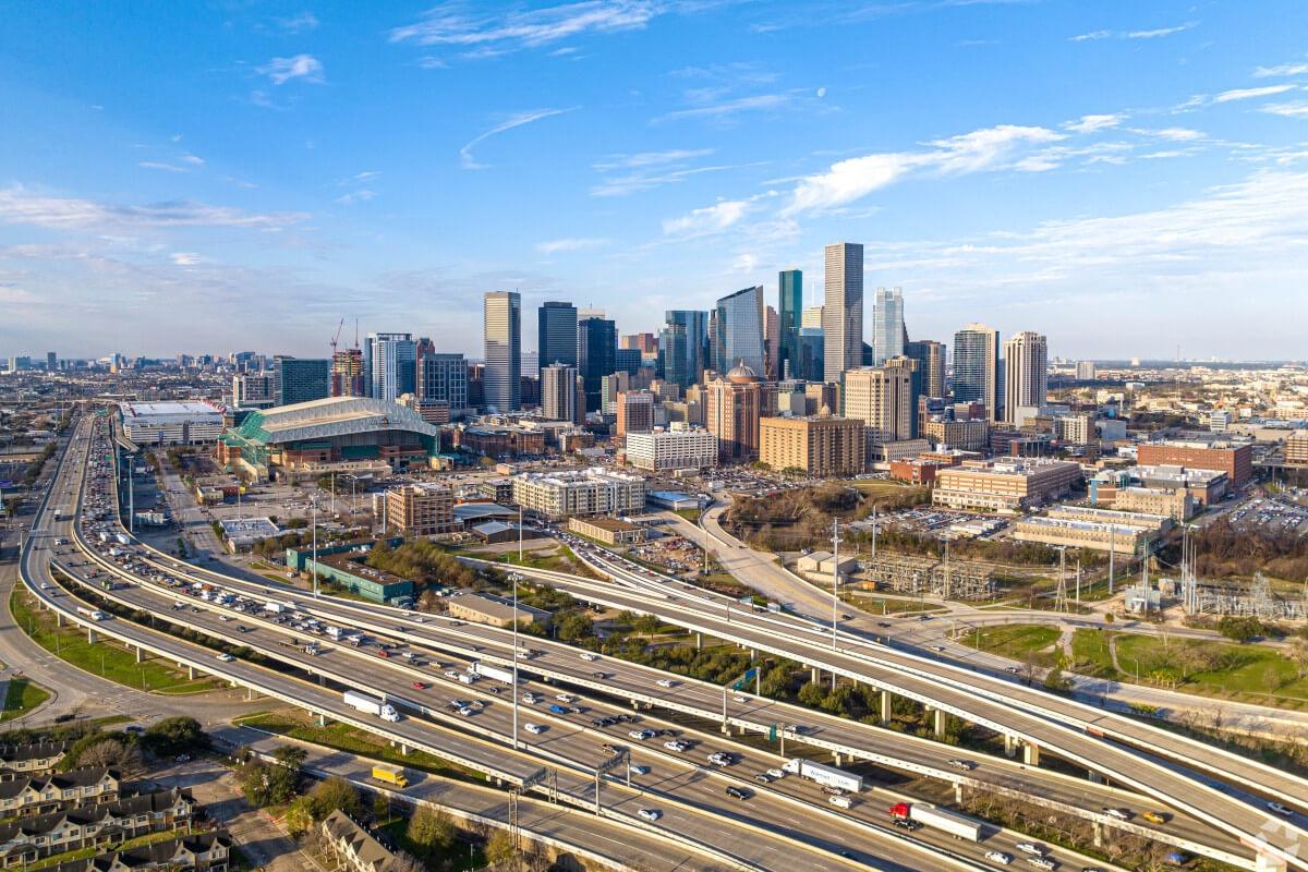 An aerial view of Downtown Houston shows skyscrapers sitting behind a highway.