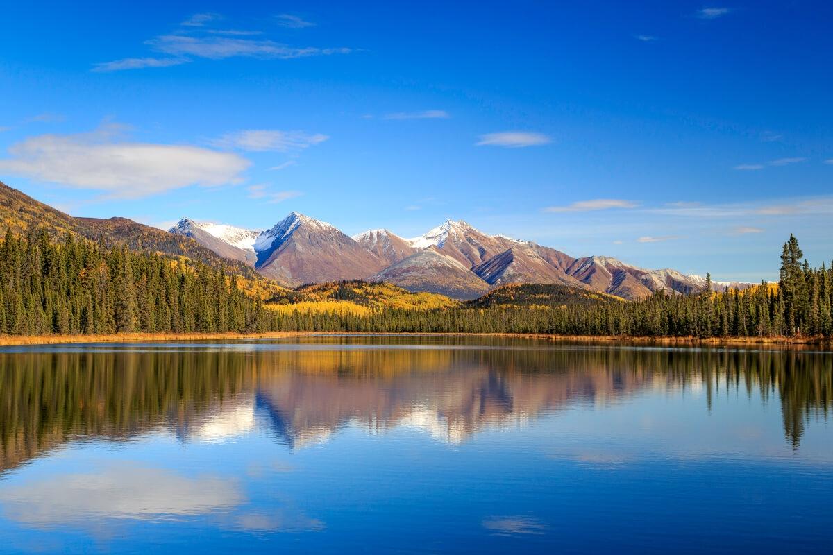 A calm lake reflecting snow-capped mountains and trees under a blue sky.