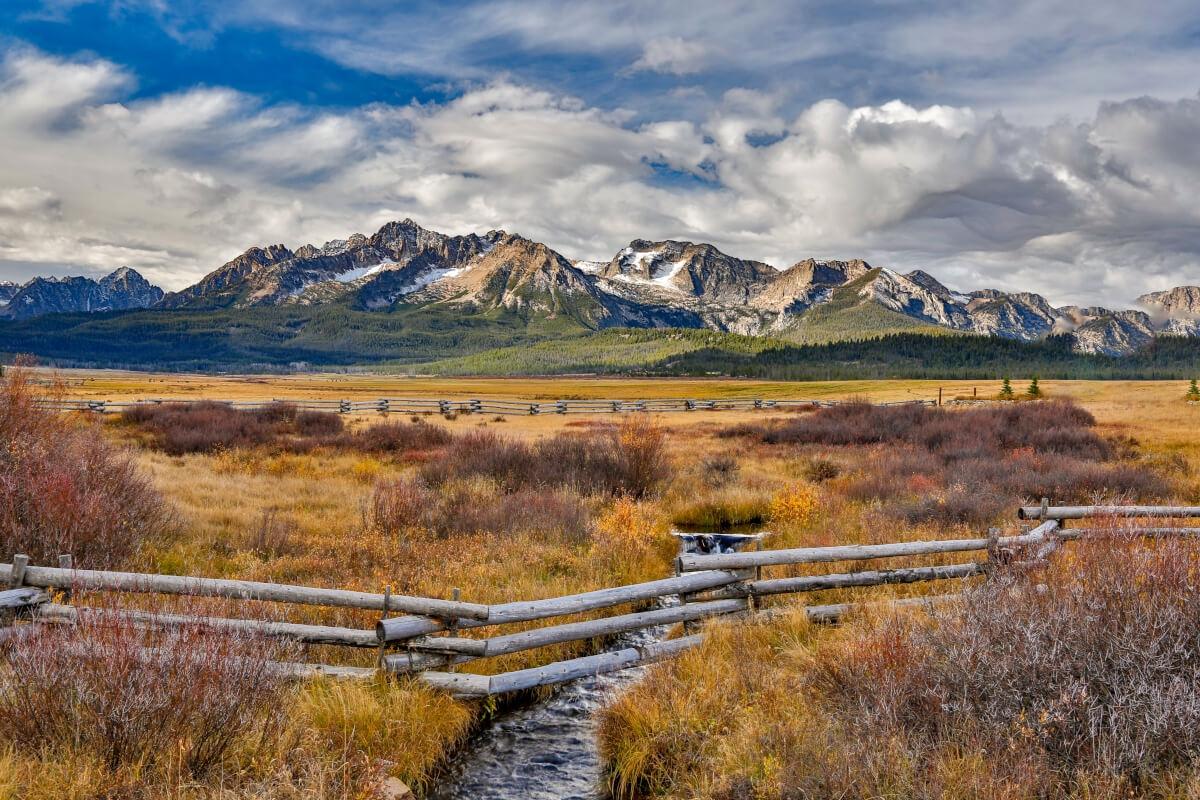 A scenic view of mountains under a blue sky with clouds, golden grassland, and a small stream with wooden fences.
