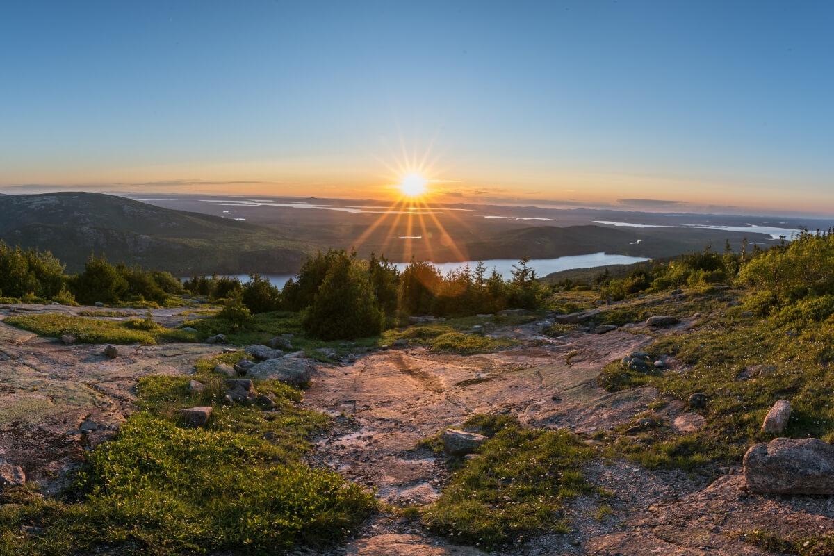 Sunrise over a beautiful landscape with rocks, green shrubs, and a lake in the distance.