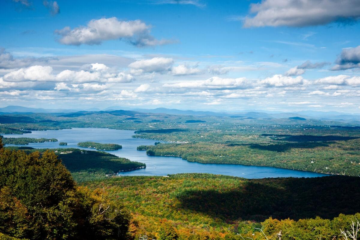A panoramic view of a lake surrounded by colorful autumn trees and clouds in the sky.