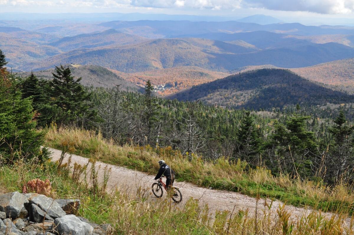 A mountain biker rides on a gravel path with colorful autumn hills in the background.