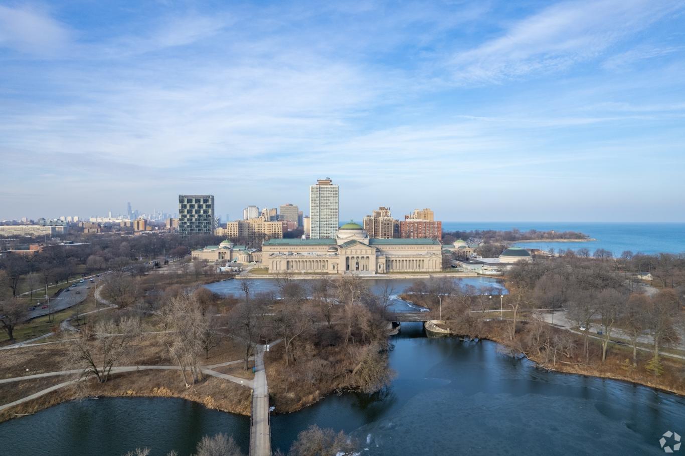 Aerial view of the Museum of Science and Industry in Hyde Park, surrounded by trees and Lake Michigan in the distance.