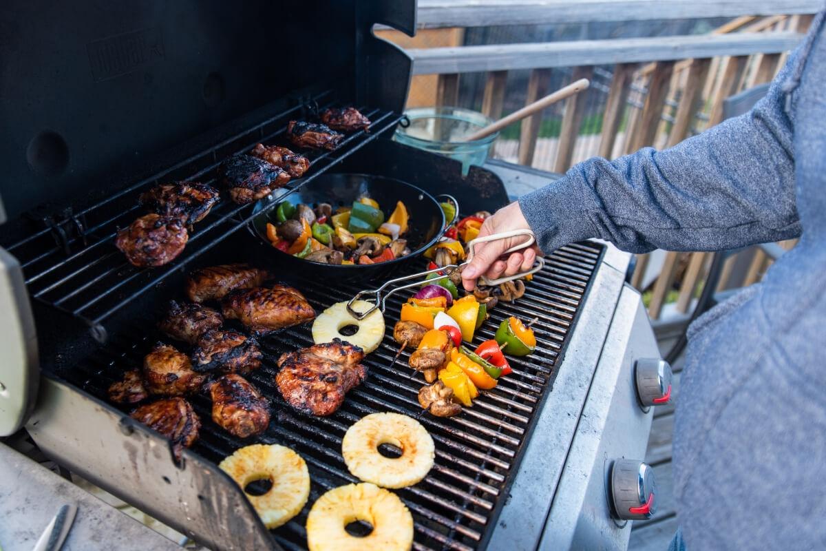 A person places pineapple rings on a grill alongside vegetable skewers and chicken thighs.