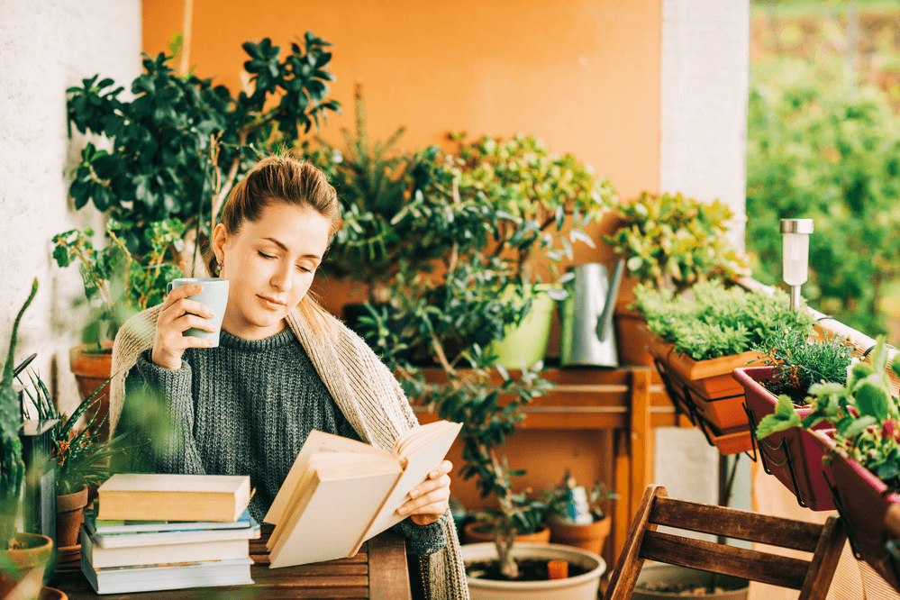 A young woman reads on a porch surrounded with plants.