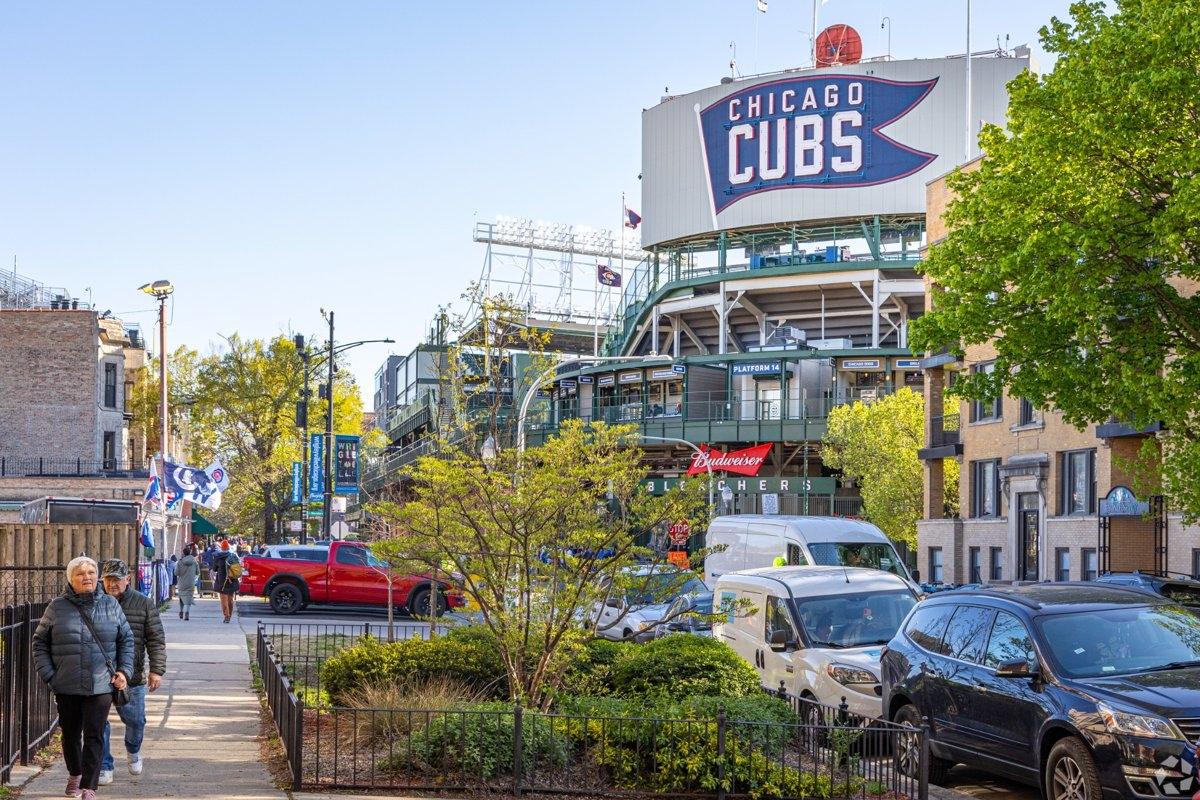 View of Wrigley Field with Chicago Cubs signage, surrounded by fans, vehicles, and nearby buildings on a sunny day.