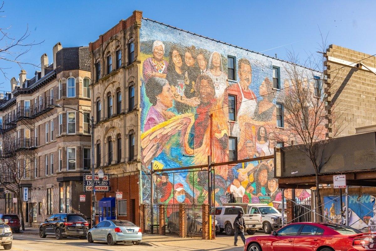 A bustling outdoor art market on a city street in Pilsen with white vendor tents and crowds of people browsing artwork.