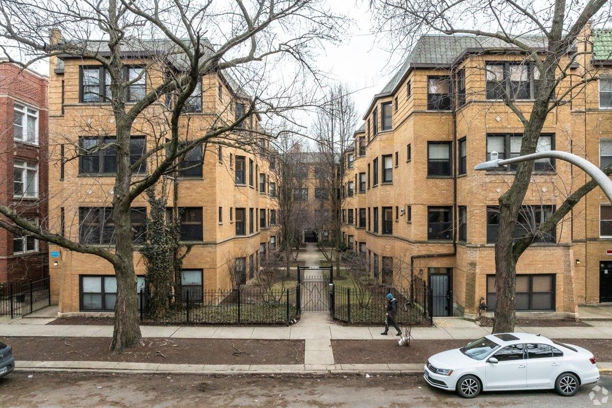 A symmetrical view of a tan-brick apartment building complex with a gated central courtyard, leafless trees lining the sidewalk, a person walking a dog past, and a white sedan parked in front.