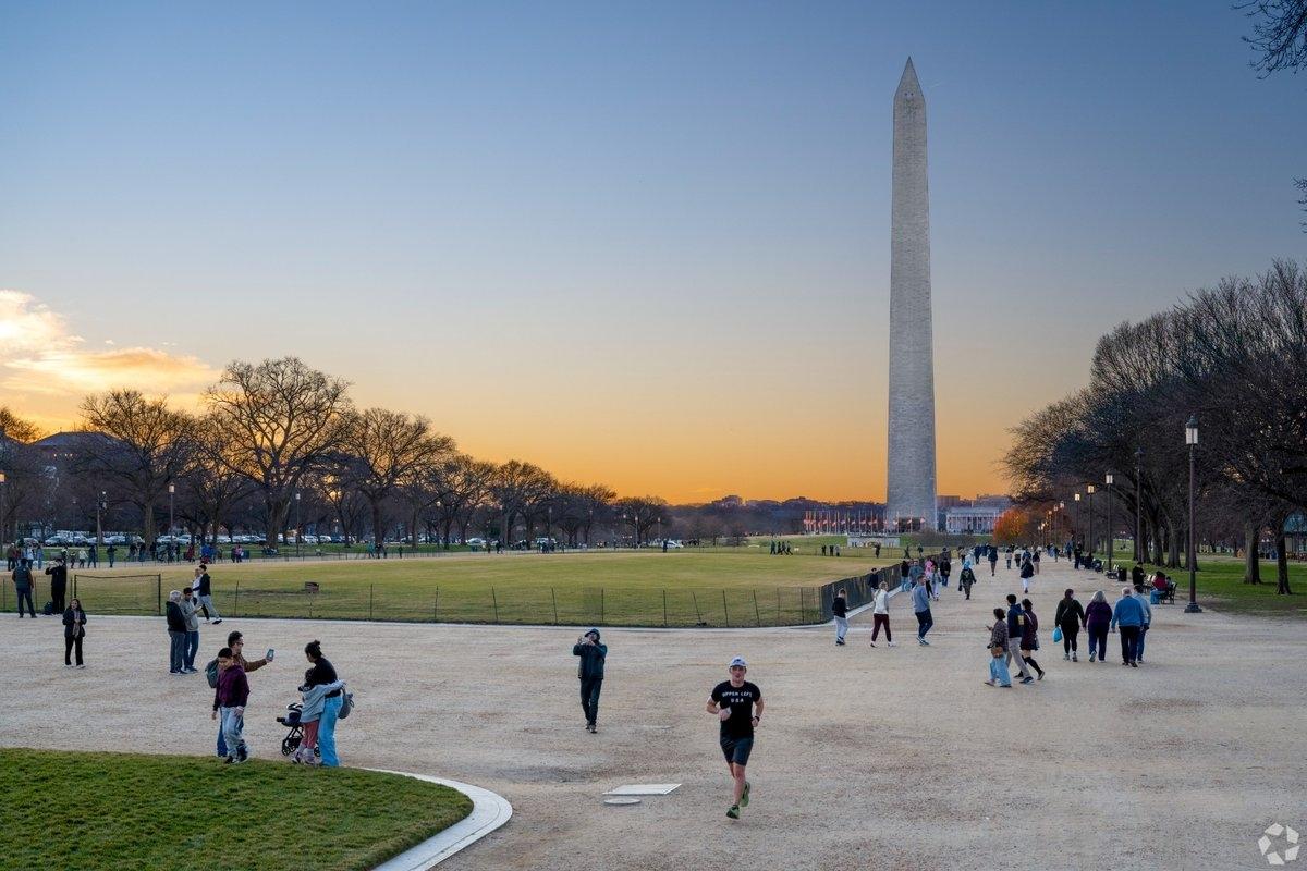 The tall Washington Monument at sunset rising above a grassy expanse with people walking and jogging on a path in the foreground.
