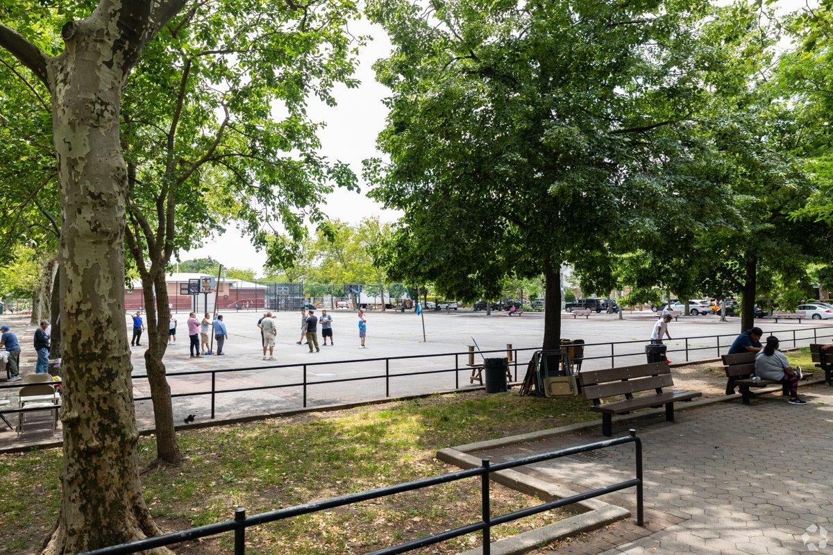 Community playground in Jackson Heights with shady trees, local families relaxing, and recreational courts ideal for outdoor apartment living.