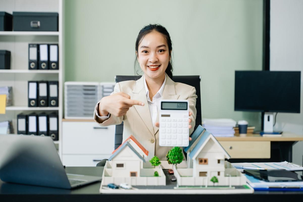 Smiling real estate agent holding a calculator and pointing at miniature houses, discussing renters insurance savings.