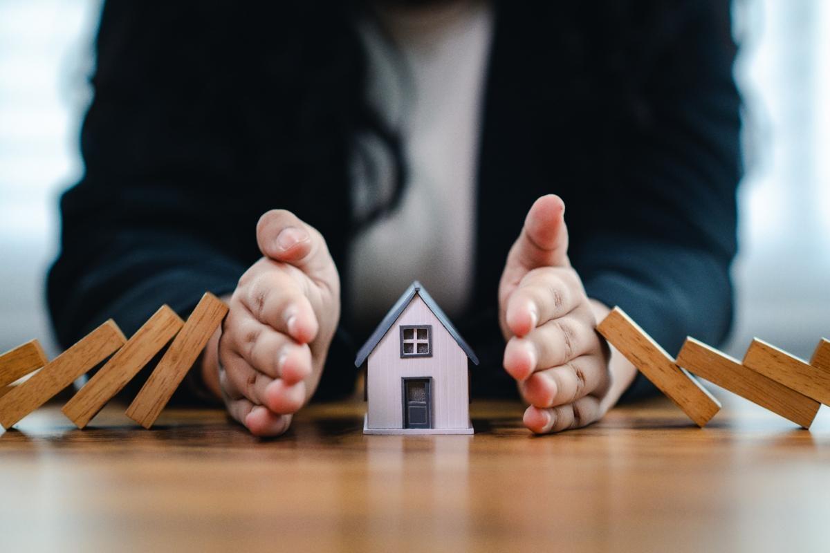 Hands shielding a small house from falling wooden blocks, symbolizing renters insurance protection coverage.