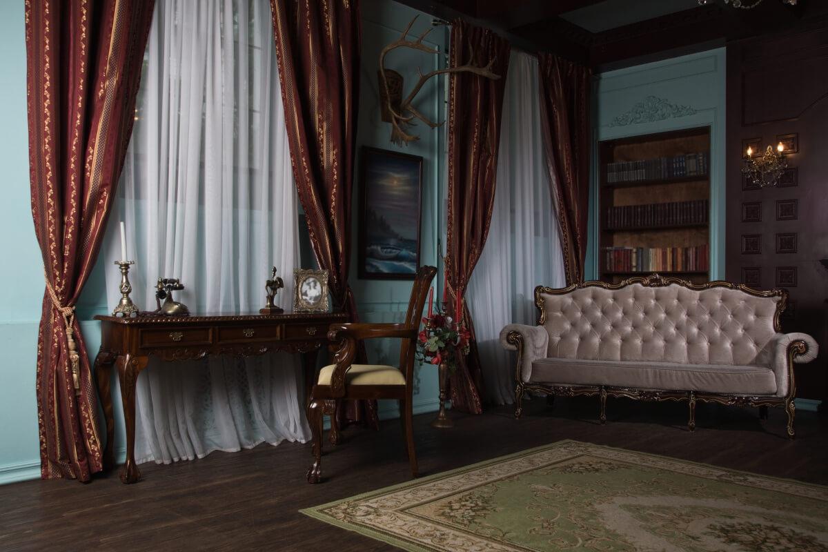A dark academia study has dark red curtains and vintage-style furniture.