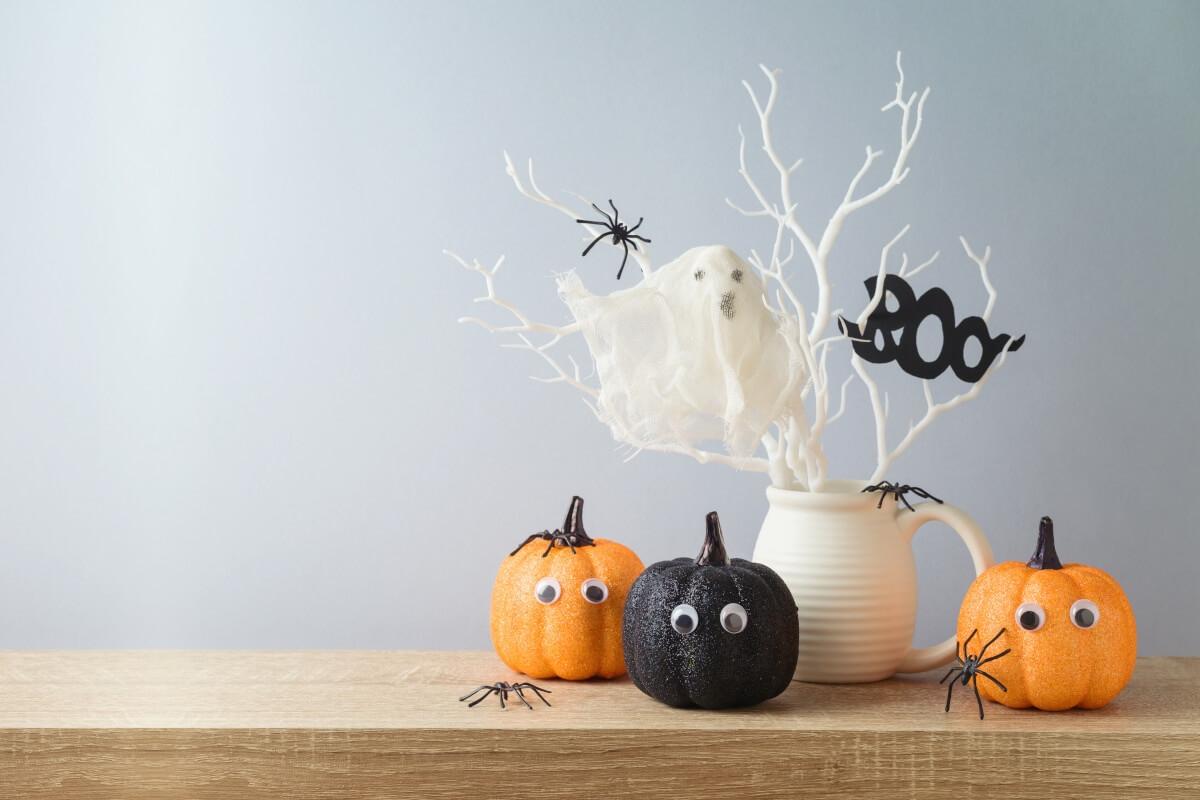 Three pumpkins with googly eyes sit on a table with plastic spiders all around.