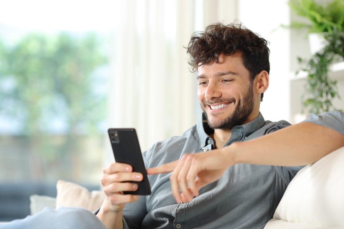 A man sitting on his sofa with a smile while using his phone