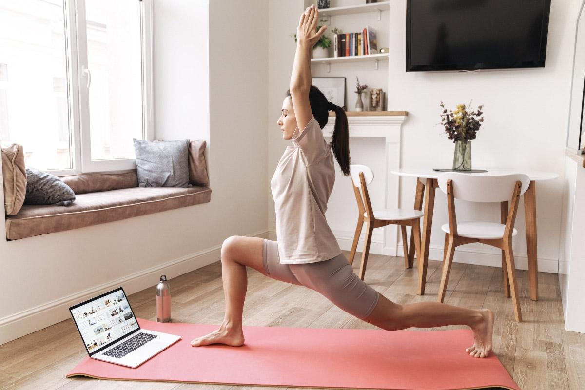 A woman does the Warrior Pose on a yoga mat in her apartment living room.