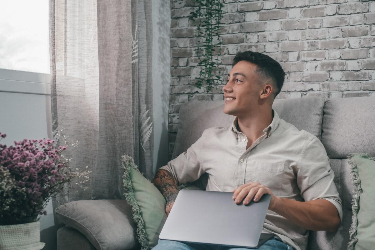 A smiling young man closes his laptop while looking out of his apartment window.