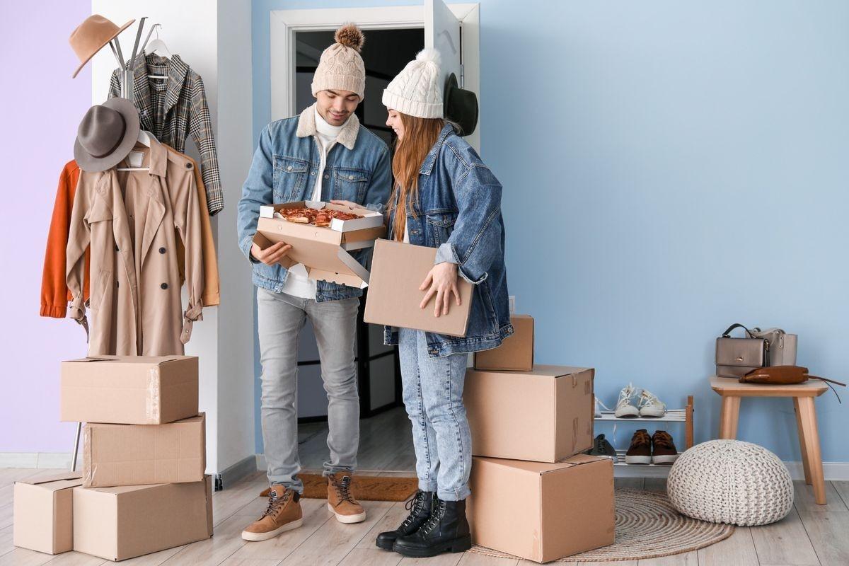 Couple wearing coats and hats, surrounded by moving boxes.
