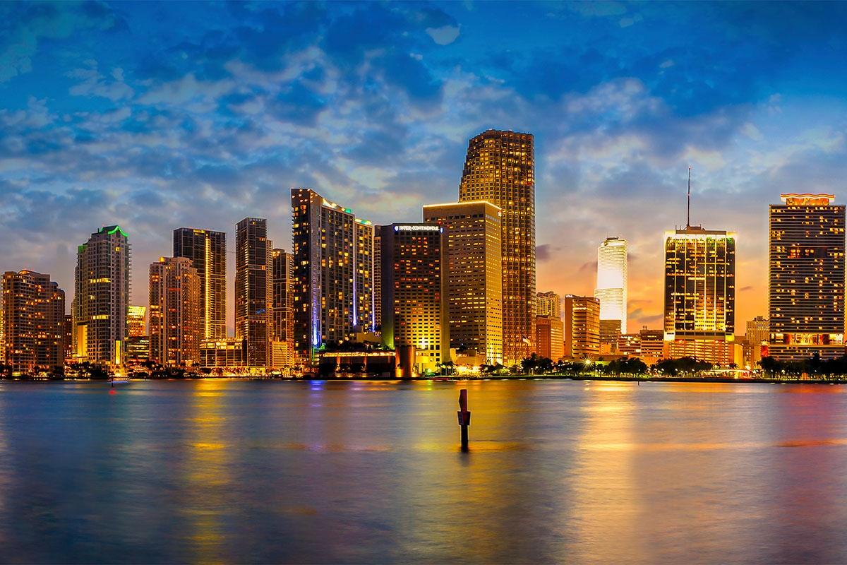 The buildings along Boston's shoreline glow with lights that reflect off the water as night falls.