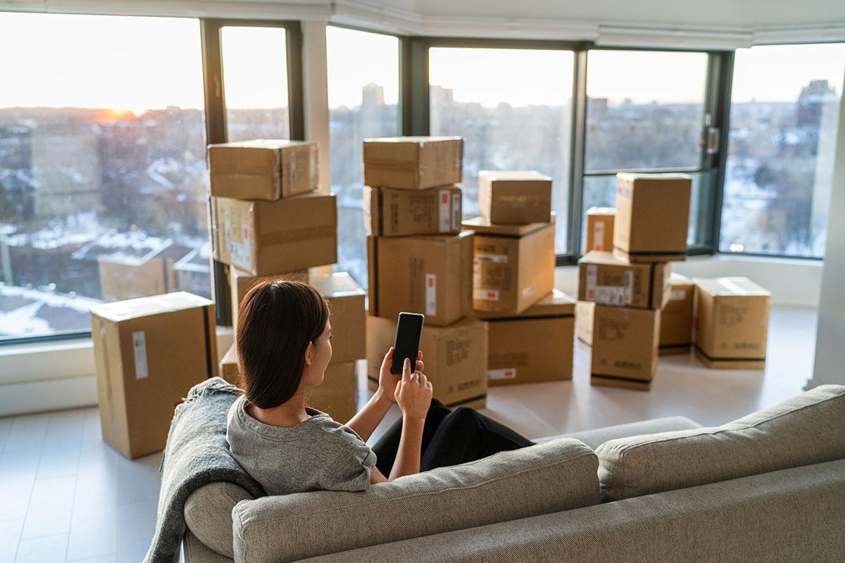 Woman looking at phone while sitting on a couch in a high-rise apartment filled with moving boxes.