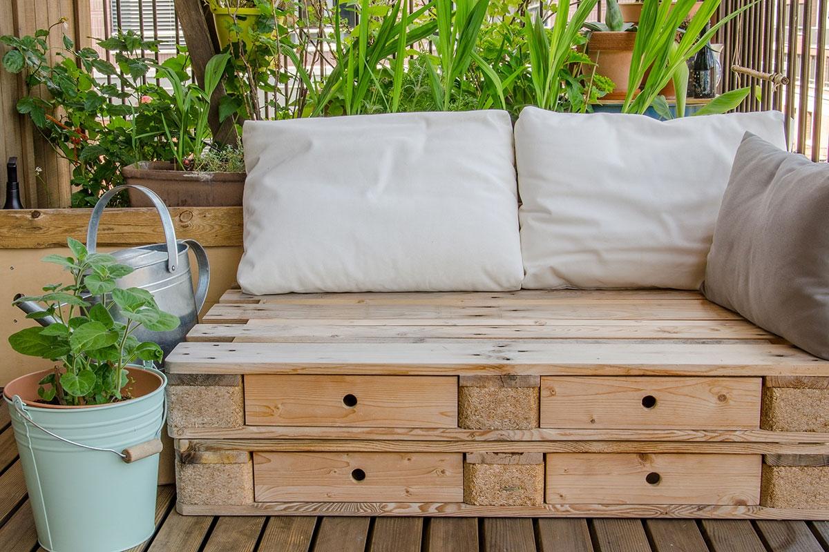 A daybed made out of pallets sits on a balcony surrounded by plants.