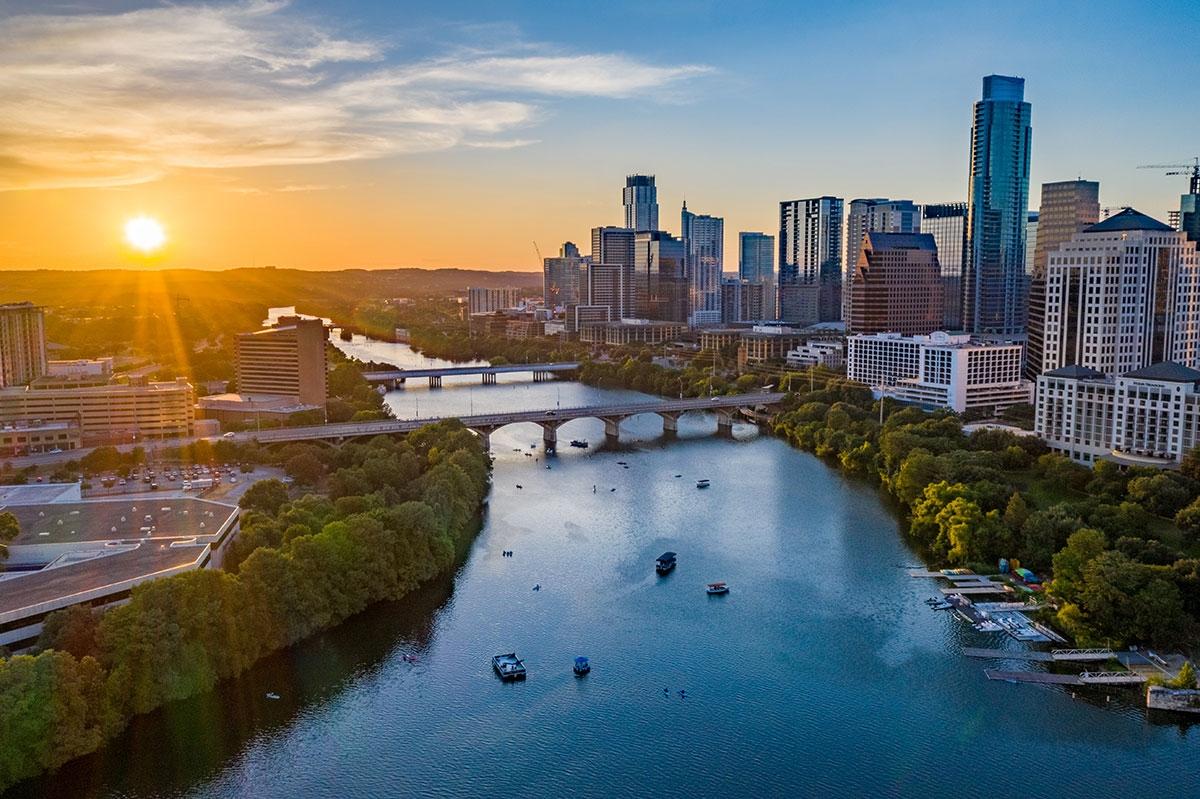 Downtown Austin at sunset, with the Colorado River flowing through the center, bridges crossing the water, kayakers visible below, and the city skyline rising in the background.