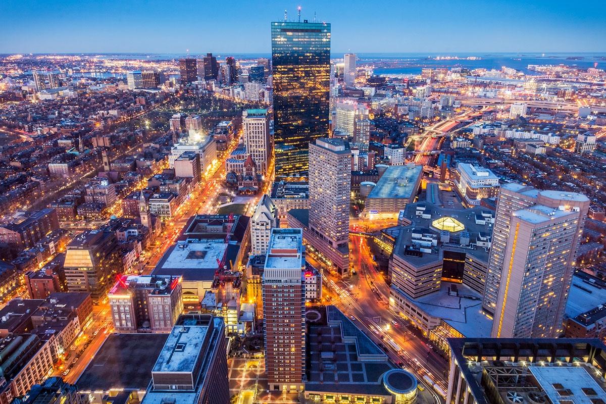 Downtown Boston is filled with illuminated skyscrapers, busy streets filled with traffic, and historic and modern buildings densely packed across the cityscape.