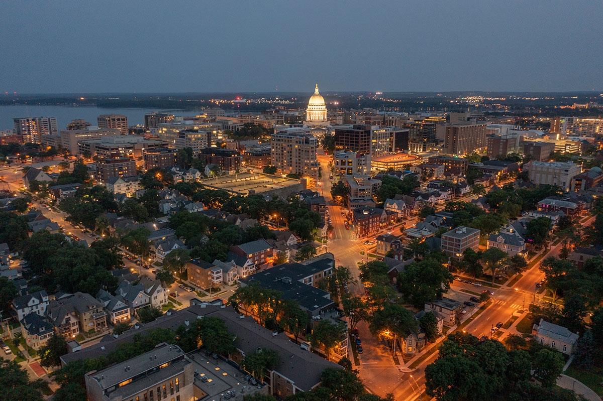 The Wisconsin State Capitol building glows in the distance, tree-lined residential neighborhoods in the foreground, and city streets illuminated by streetlights.
