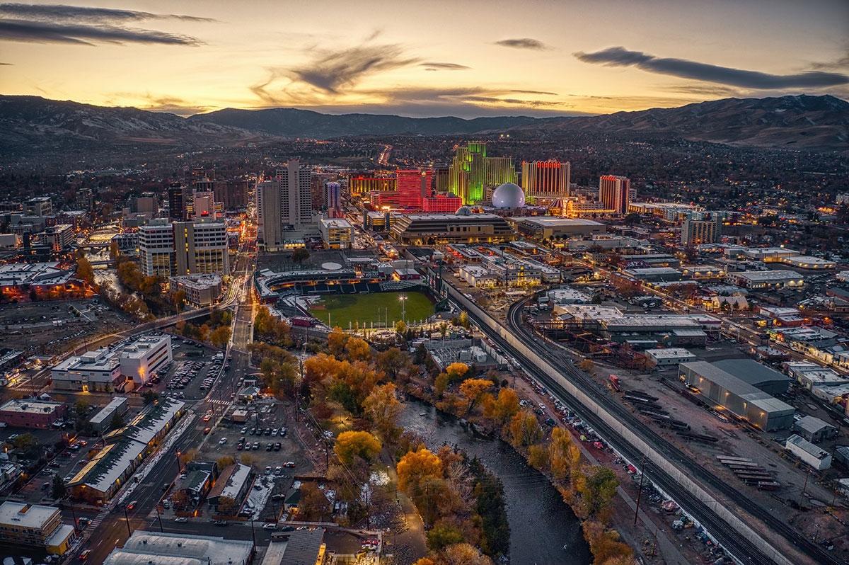 Reno contains brightly lit casinos and hotels, a baseball stadium, rail lines, and surrounding neighborhoods with mountains visible in the distance.