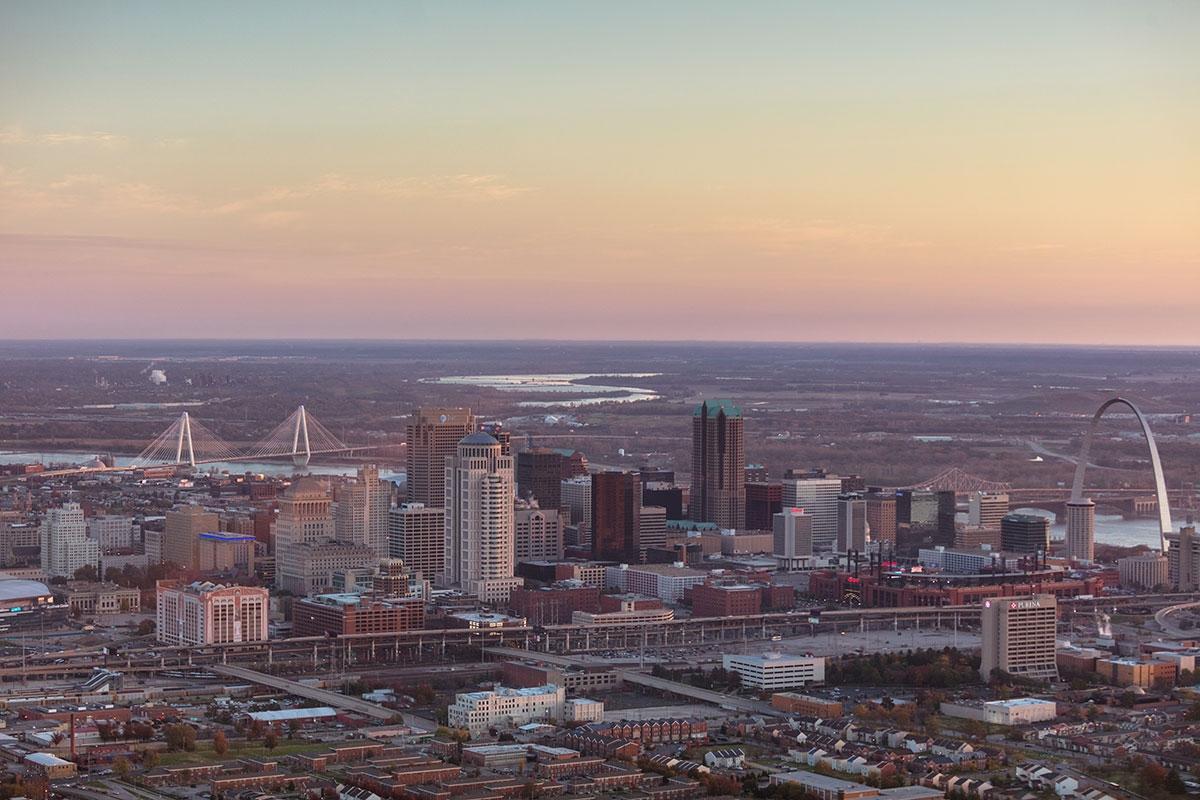 St. Louis with the Gateway Arch visible on the right, the Mississippi River beyond the skyline, and highways and city buildings spread across the foreground at dusk.