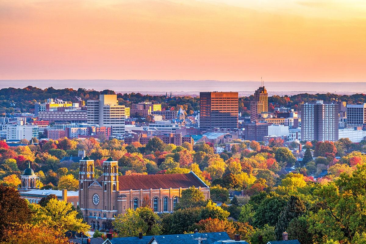 Downtown Syracuse during autumn, featuring mid-rise buildings surrounded by colorful fall foliage and a church with towers in the foreground under a warm evening sky.