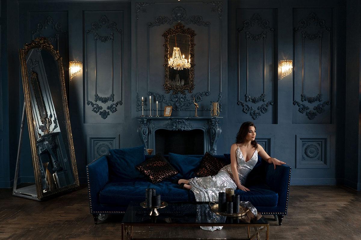 A woman in an evening gown reclines on a velvet sofa in a richly decorated room with mirrors, candles, and deep blue walls.