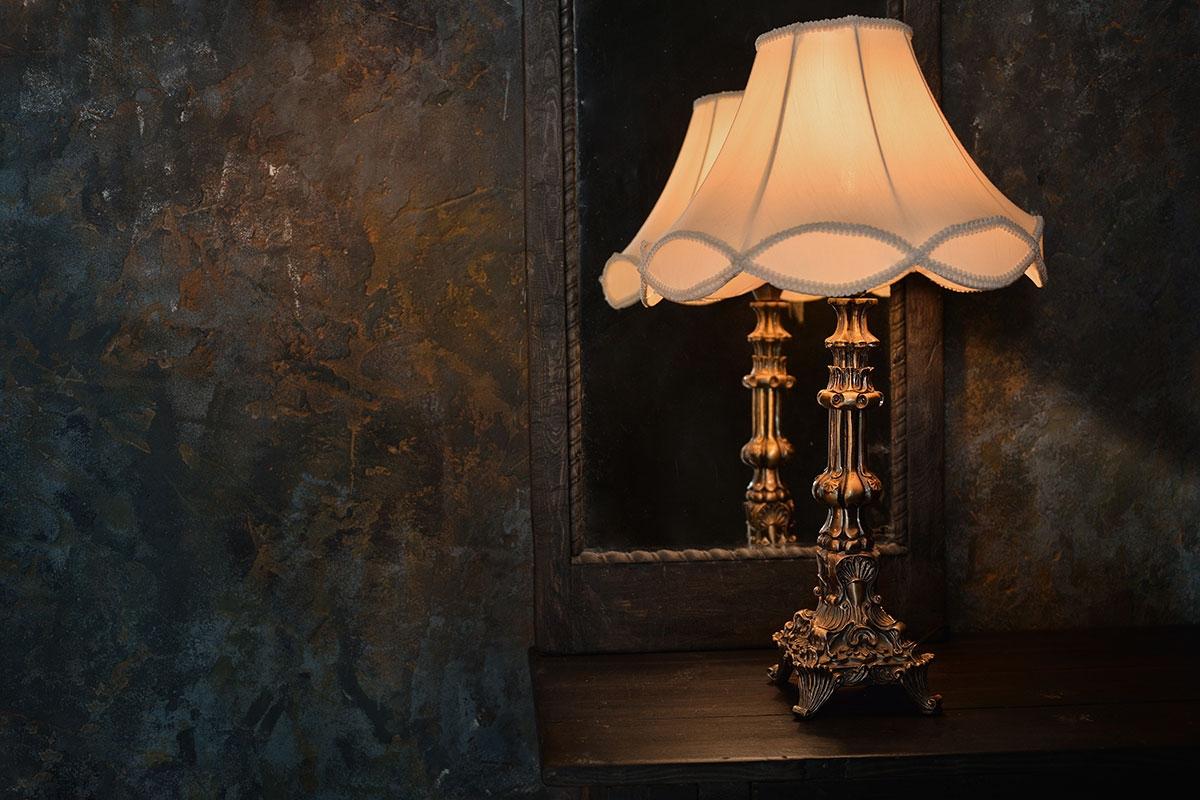 An ornate vintage table lamp with a fabric shade casts warm light against a dark, textured wall, emphasizing a vamp romantic design..