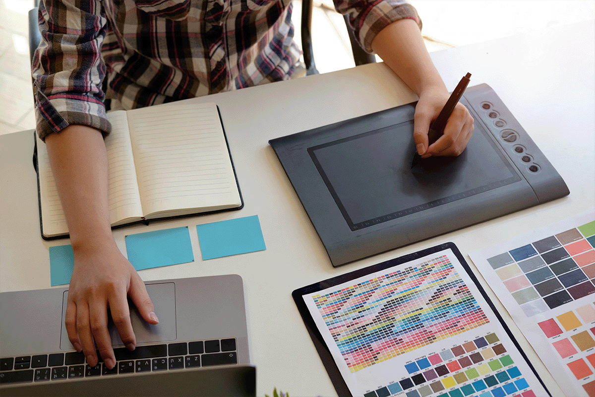 Digital artist using drawing tablet with a workspace featuring a notebook, sticky notes, color palettes, and a MacBook