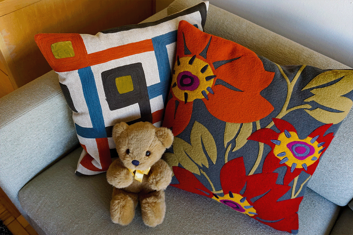 Little brown teddy bear in front of two throw pillows. One is blue, red, and orange and depicts flowers. The other is white with geometric patterns.