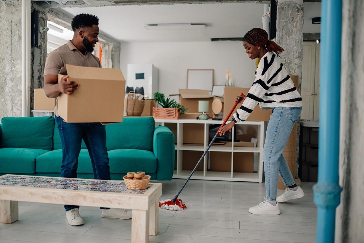 Woman mopping living room while man carries moving boxes.