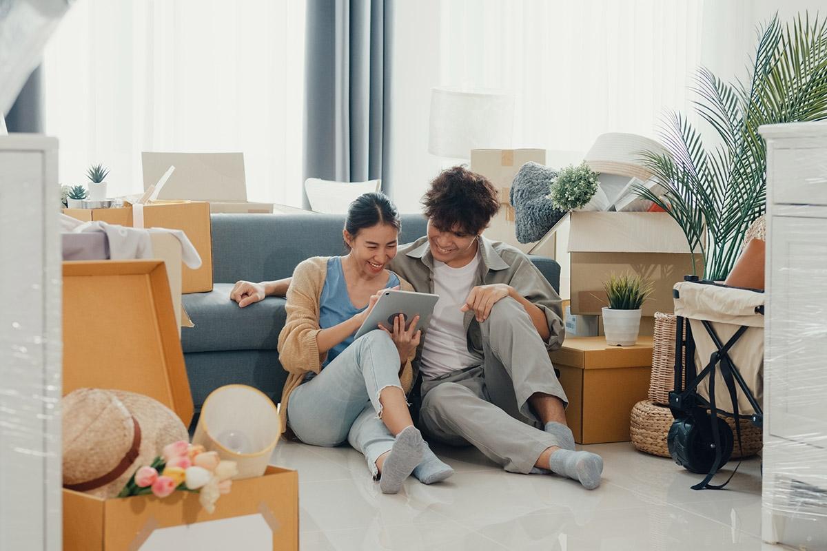 Smiling man and woman sitting on the ground, looking at a tablet as they are surrounded by moving boxes.
