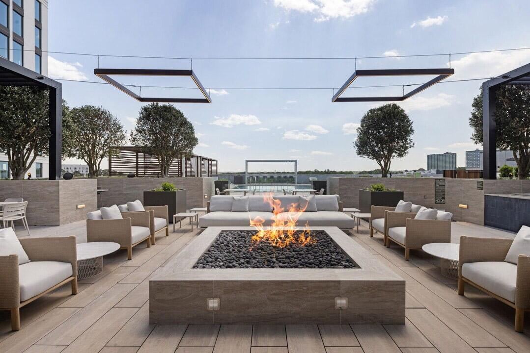 An outdoor firepit sits surrounded by couches with a pool in the background atop the roof a luxury apartment community.