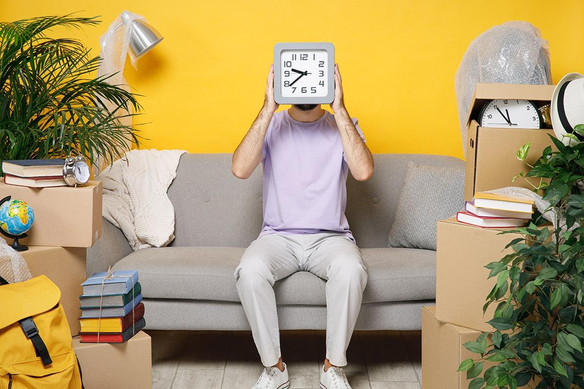 Renter sitting on a couch surrounded by moving boxes and holding a clock in front of their face.