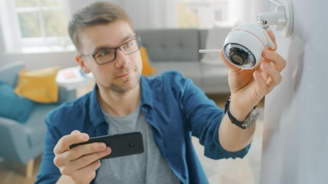 Man adjusts a security camera on a wall while holding his smartphone.