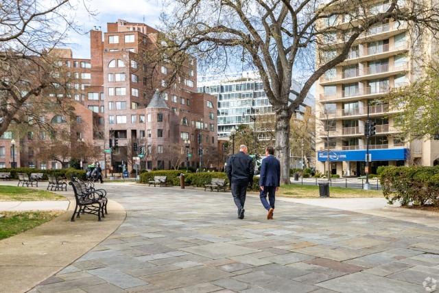 Two businessmen walking on a path through a park with apartment buildings in the background.