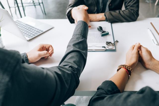 Two people shaking hands over contract with someone sitting next to them.