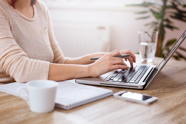 Woman holding pen and typing on laptop with notebook and phone next to her.