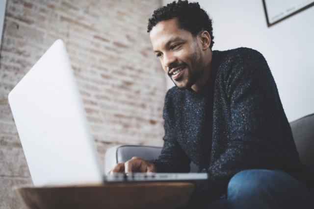 A smiling young man searches for apartments on a laptop.