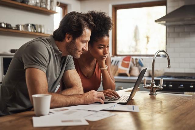 A couple sitting in their kitchen and browsing on a laptop. The man is wearing a grey T-shirt. The woman has her hair tied up and is wearing an orange T-shirt.