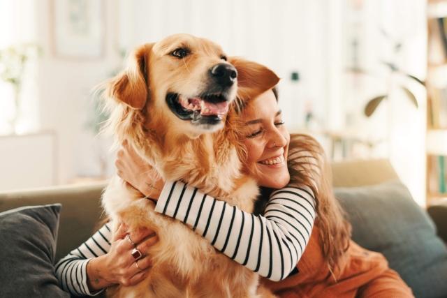A woman with long brown hair in a striped shirt and orange overalls hugs her Golden Retriever.