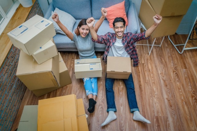 Man and woman sitting on the floor against a couch looking victorious with their arms in the air as they are surrounded by moving boxes.