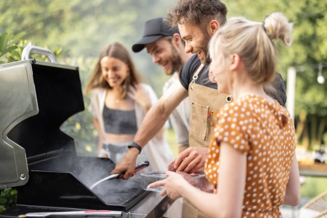 Three young people smile as their friend flips a burger on a grill.