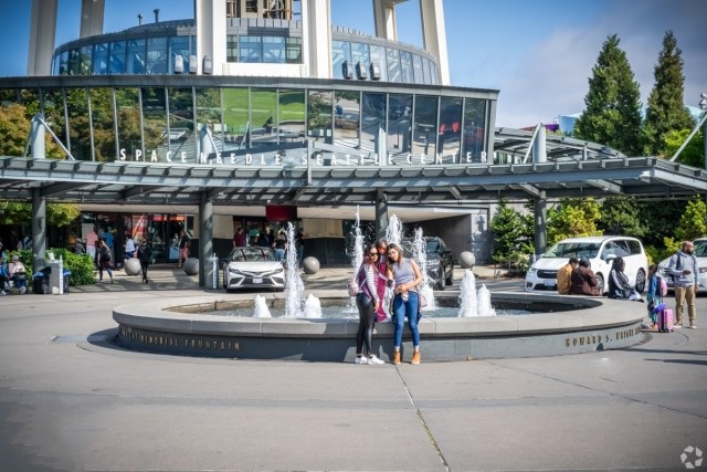 Three women pose in front of a circular fountain with water jets at the base of the Space Needle in Seattle, surrounded by visitors and parked cars.